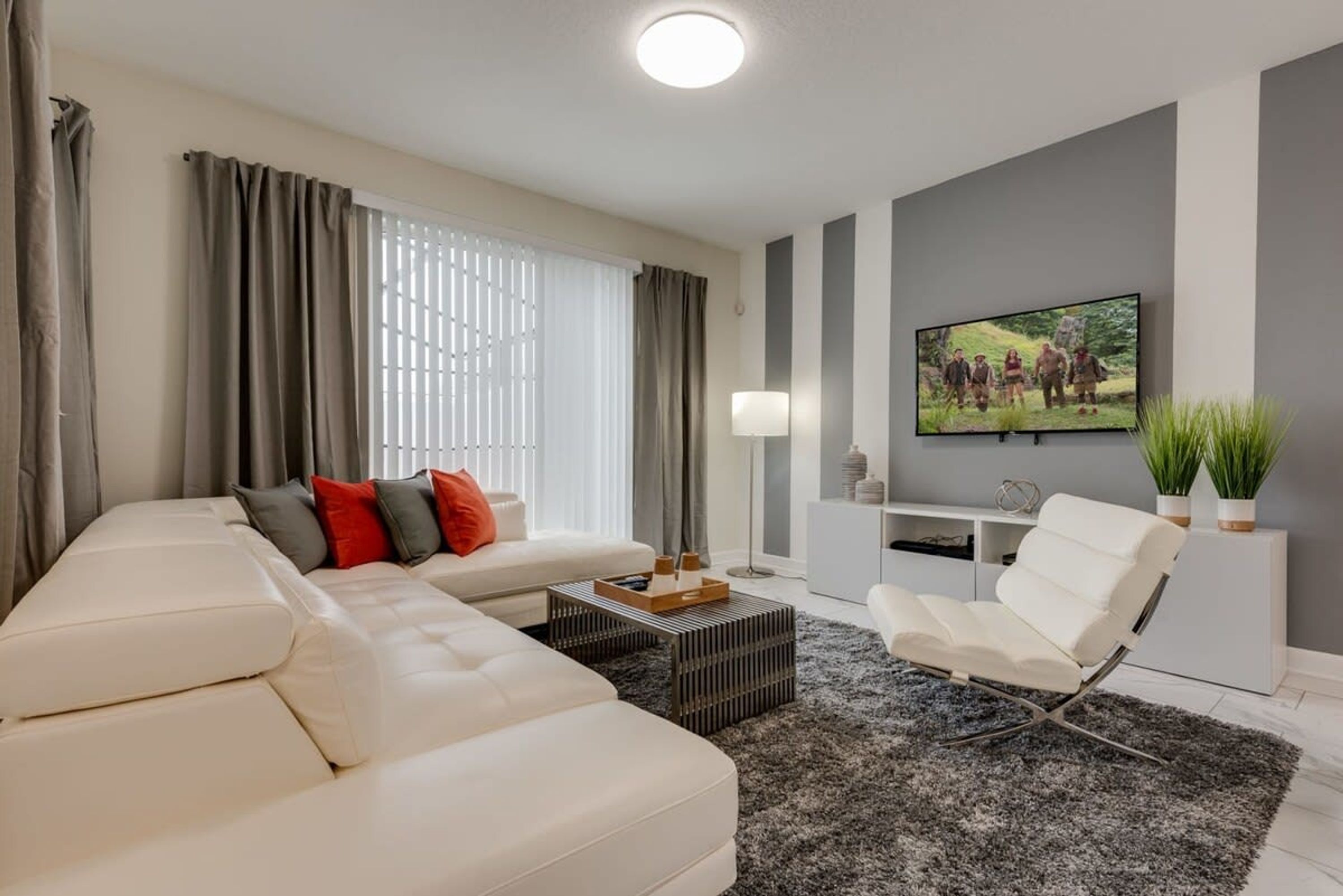 A modern living room with a white sofa, a coffee table, a white armchair, and a flat-screen TV mounted on the wall.