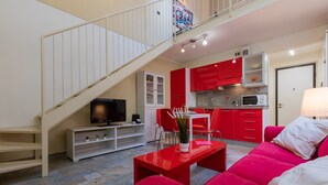 Una sala moderna con un sofá rojo, una mesa de centro y un televisor. Una escalera conduce a un balcón. La cocina tiene armarios rojos y un microondas.