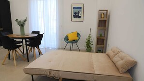 Ett modernt rum med en säng, ett matbord med stolar, en liten stol med en gul kudde, en bokhylla och en krukväxt.