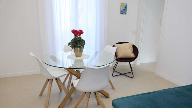 Ett modernt matområde med ett glasbord och vita stolar, en krukväxt och en soffa i turkos.