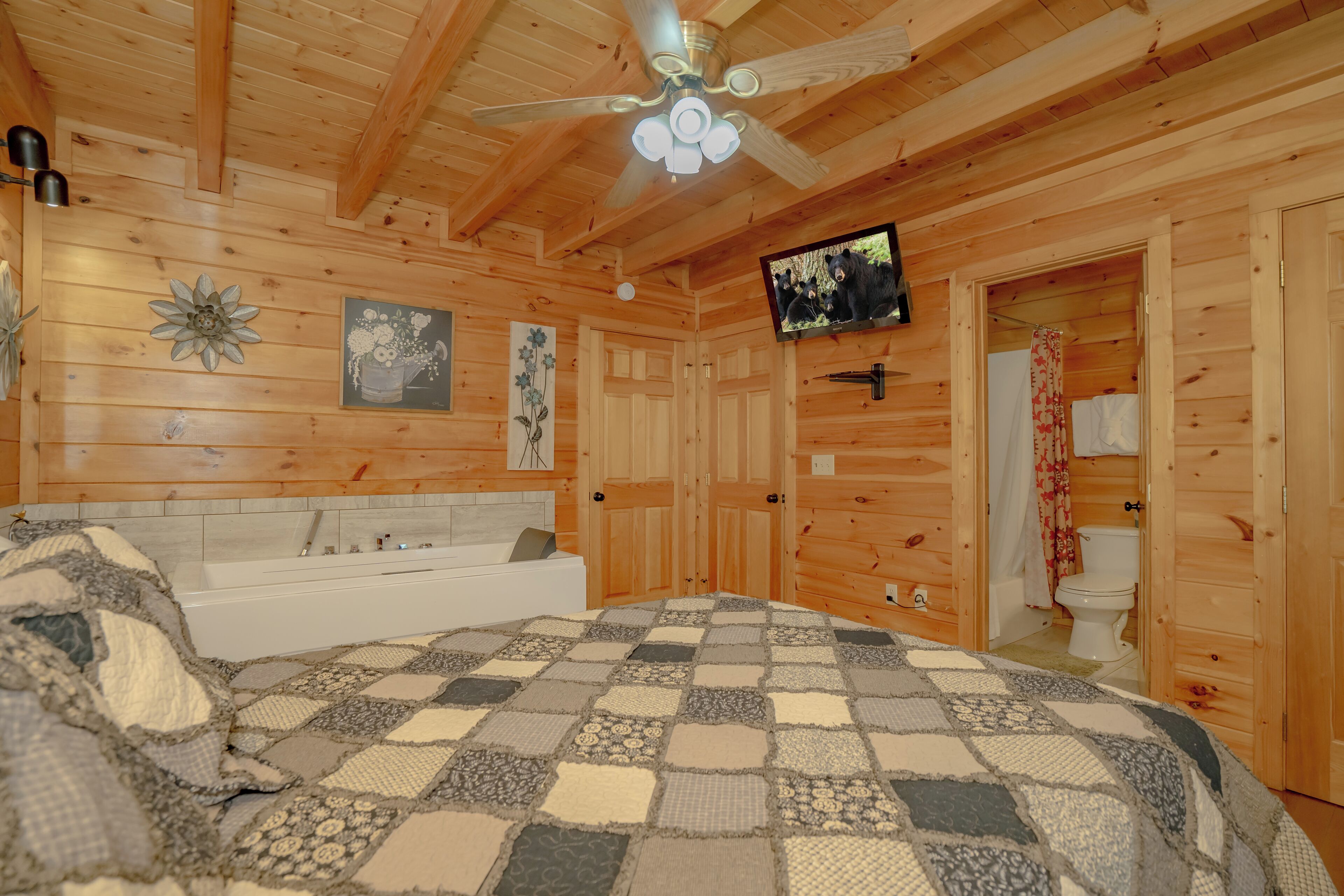 A wooden cabin bedroom with a checkered bedspread, a ceiling fan, a mounted TV, and a bathtub.