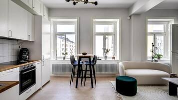 Una cocina moderna con armarios blancos, una mesa de comedor con sillas, una cafetera y un radiador. Hay un sofá y un reposapiés verde en la sala de estar.