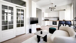Una sala moderna con un televisor de pantalla plana, un sofá y una mesa de centro. Se aprecia una cocina al fondo con armarios blancos y una encimera de madera.