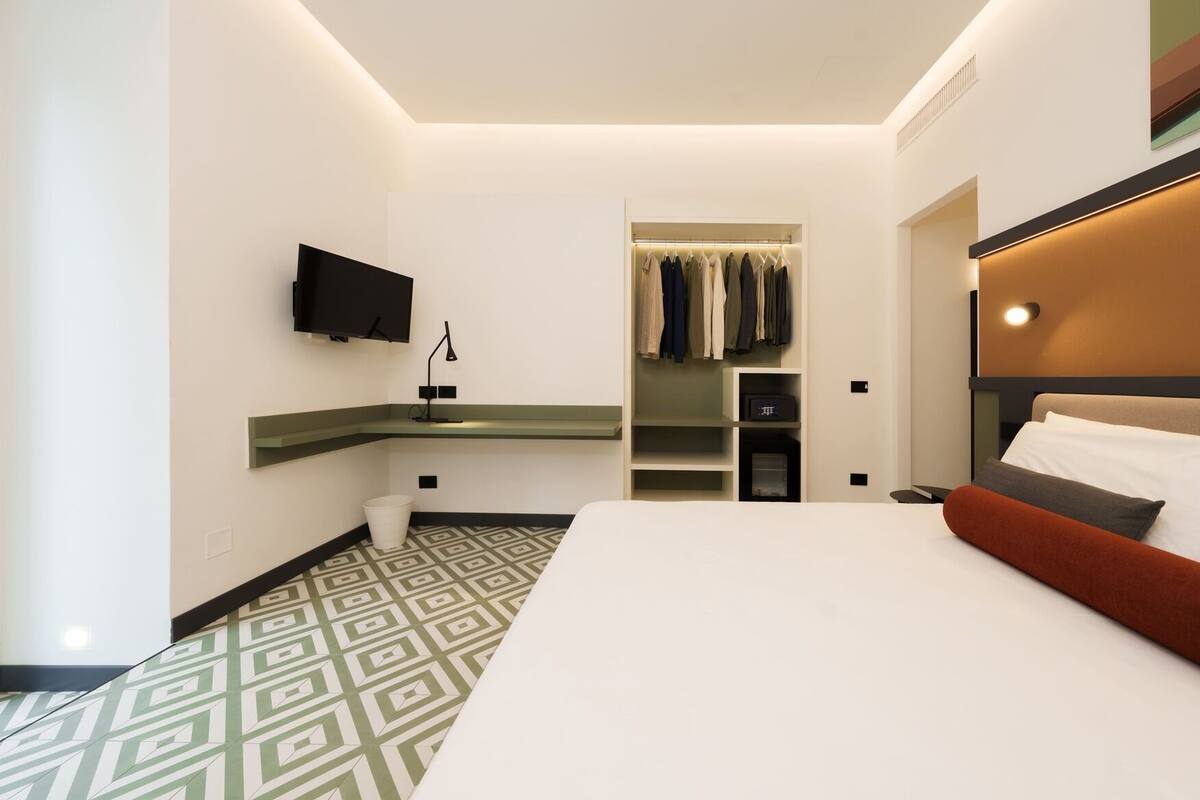 A hotel room with a bed, a TV mounted on the wall, a closet with hanging clothes, and a bedside table.