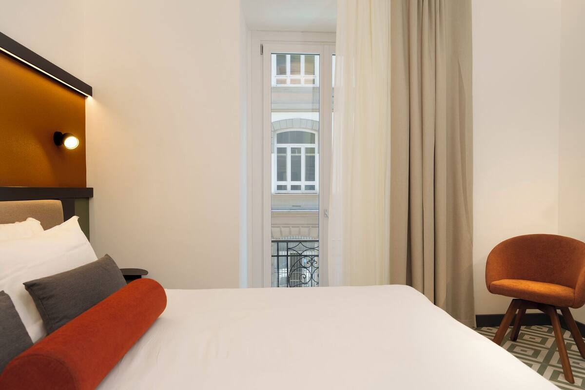 A hotel room with a bed, a chair, a window with curtains, and a view of a building.