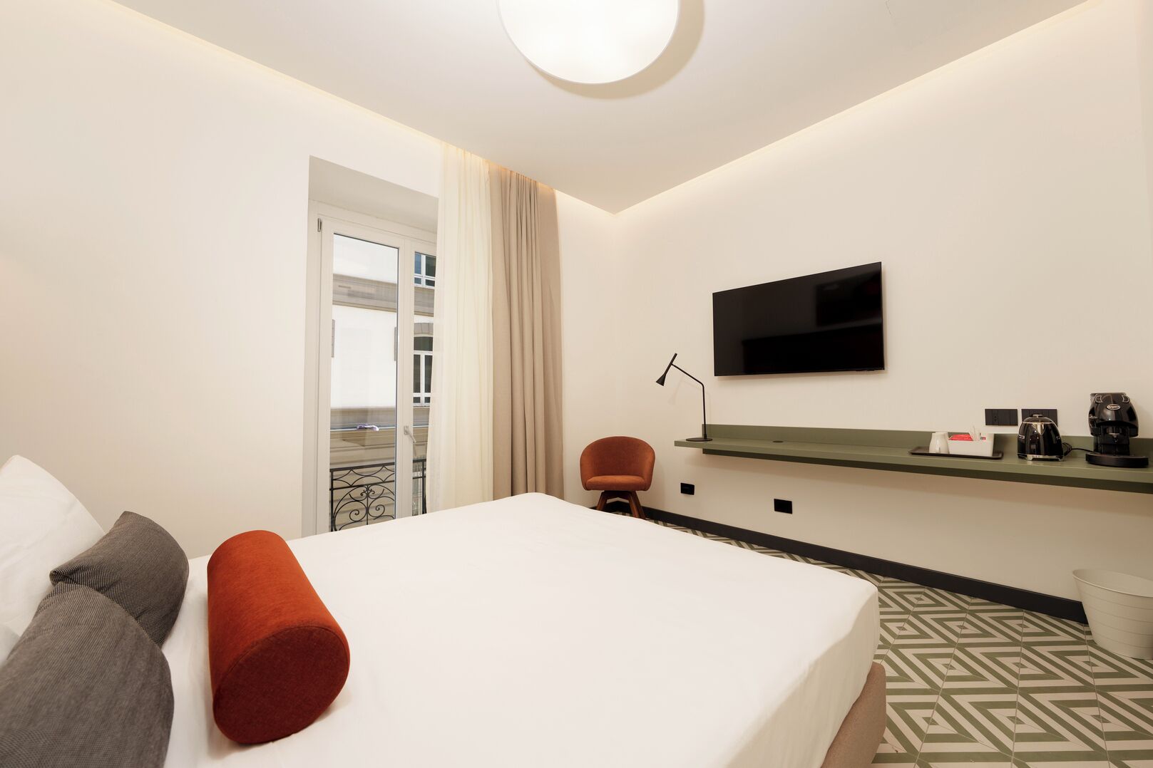 A hotel room with a bed, a chair, a TV, a coffee machine, and a small shelf.