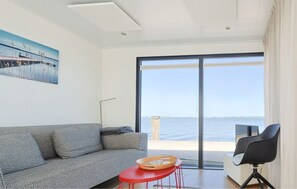 A modern room with a sofa, a small red table, and a chair. It has a large window with a view of the sea and a bridge. - Earrebarre Houseboat - Overnight stay on the water (Offingawier)