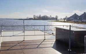 A wooden deck with a metal railing overlooking a body of water, a hot tub, and houses in the background. - Earrebarre Houseboat - Overnight stay on the water (Offingawier)