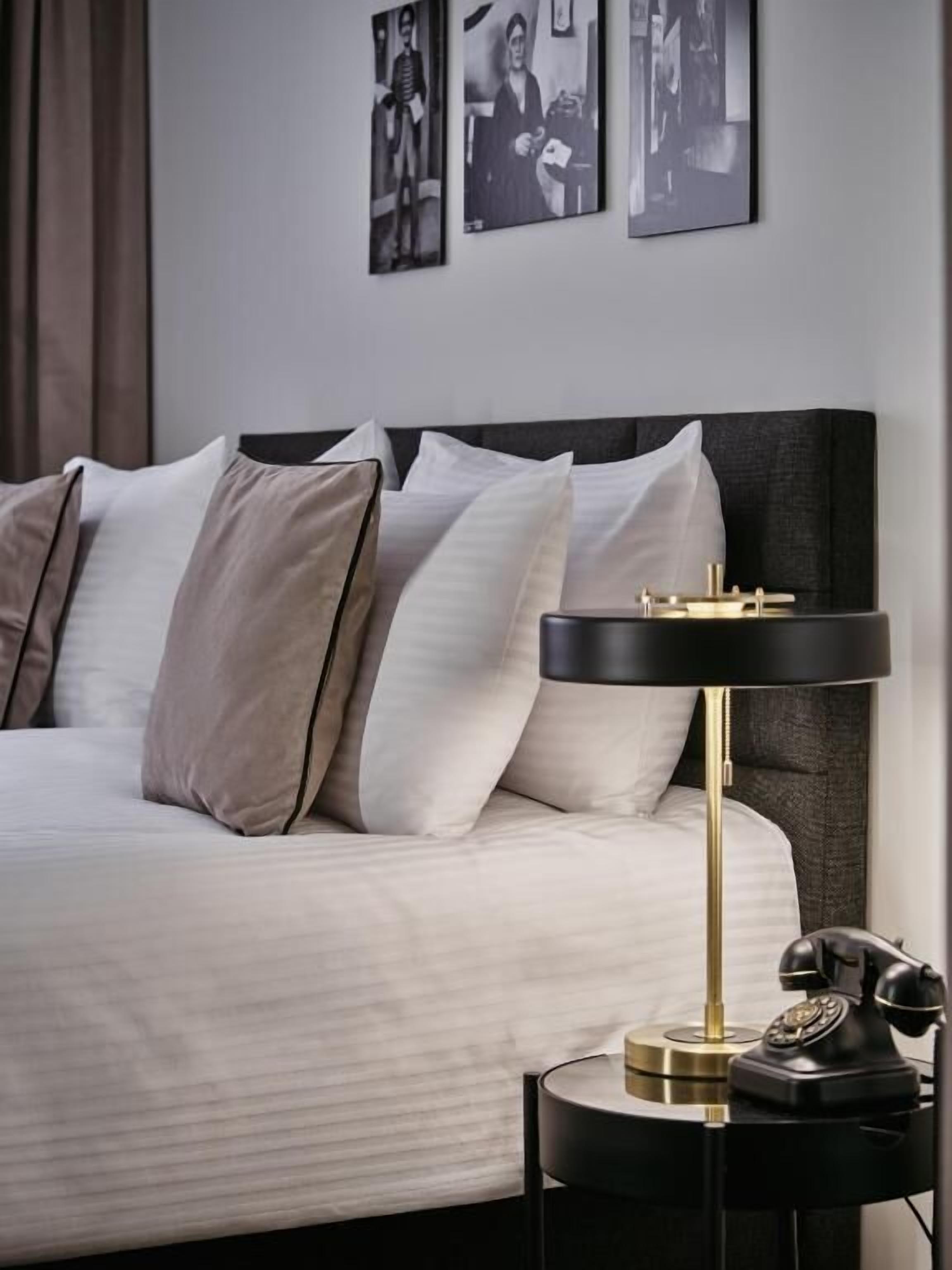 A neatly made bed with a modern headboard, a bedside table with a lamp and a telephone, and framed pictures on the wall.