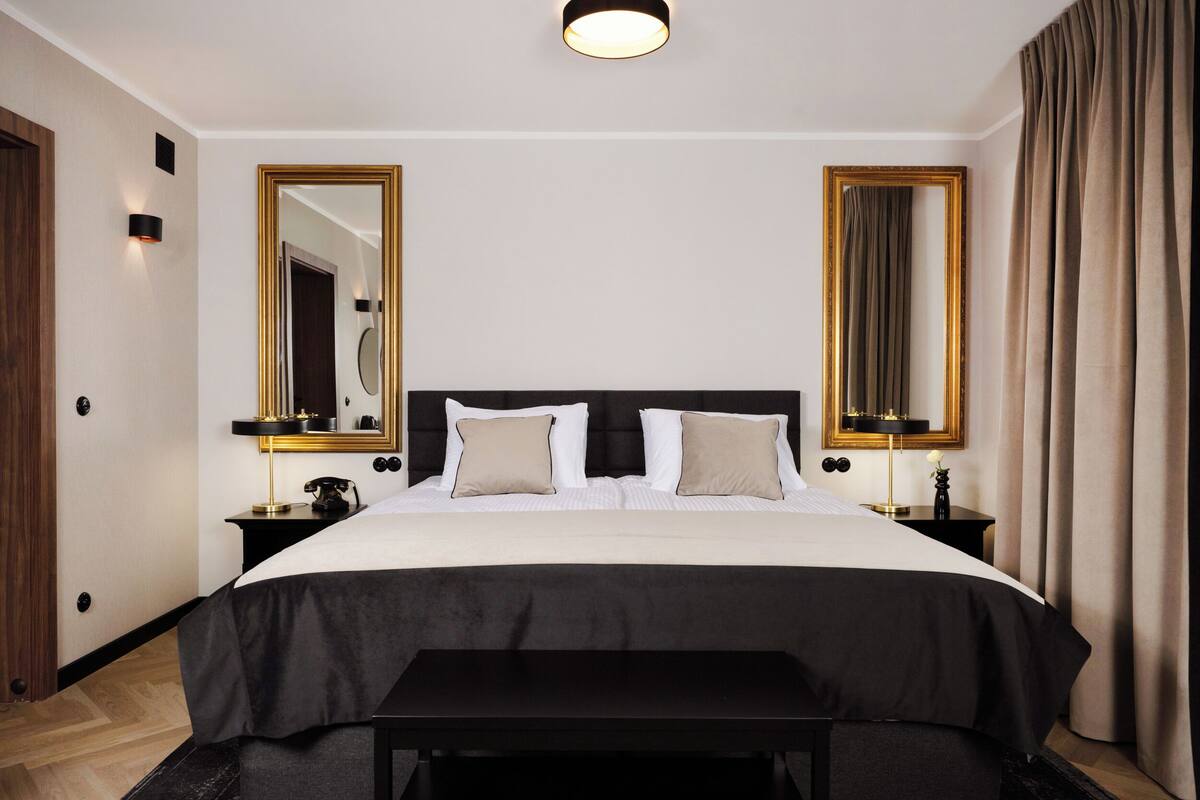 Una habitación de hotel con una cama grande, dos mesitas de noche, un banco y dos espejos amplios.