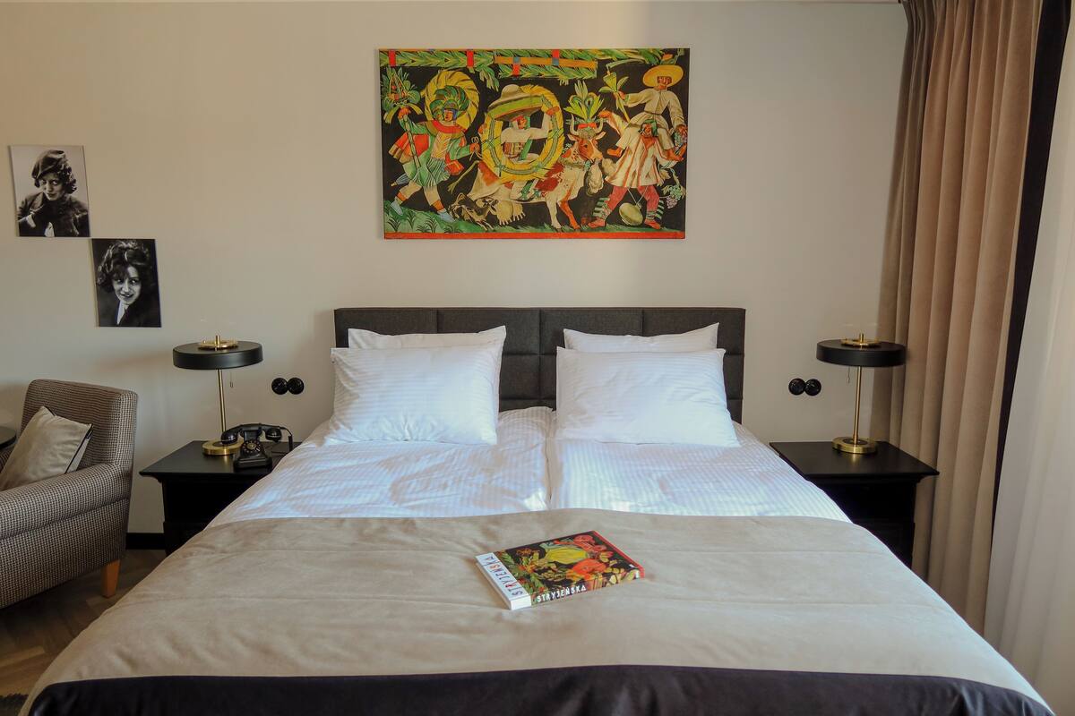 Una habitación de hotel con una cama grande, dos mesitas de noche, una silla y un cuadro en la pared.