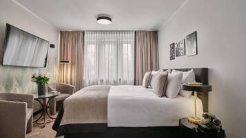 A modern hotel room with a large bed, a desk, a chair, a TV mounted on the wall, and a window with curtains.
