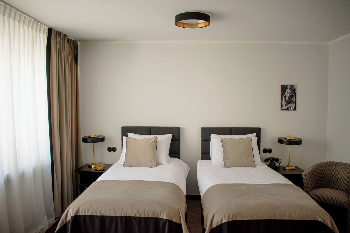 Habitación de hotel con dos camas, una mesita de noche con lámpara, una silla y un cuadro en la pared.