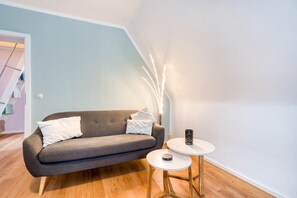 Un salon moderne avec un canapé gris, deux coussins blancs, une petite table blanche sur laquelle se trouve un objet noir, et un lampadaire fin et élancé.