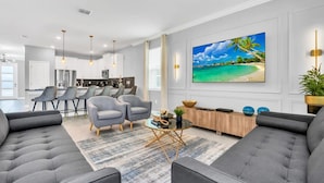 A modern living room with a grey sofa, a coffee table, and a TV mounted on the wall. There is a kitchen area in the background with bar stools and a dining table. - Luxury Incredible Themed Home, best vacation (Kissimmee)