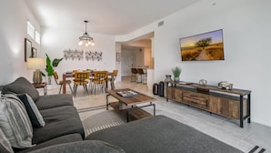 A modern living room with a sofa, coffee table, and dining area with chairs. A TV is mounted on the wall, and there is a plant on the side. - Spacious, Beautiful 3-BR Condo, Prime Location (Kissimmee)