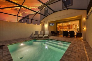 Ein Swimmingpool mit Blick auf einen Sonnenuntergang, der durch eine geometrische Glasstruktur sichtbar ist, eine Terrasse mit Stühlen sowie ein Einblick in eine Küche und einen Essbereich.