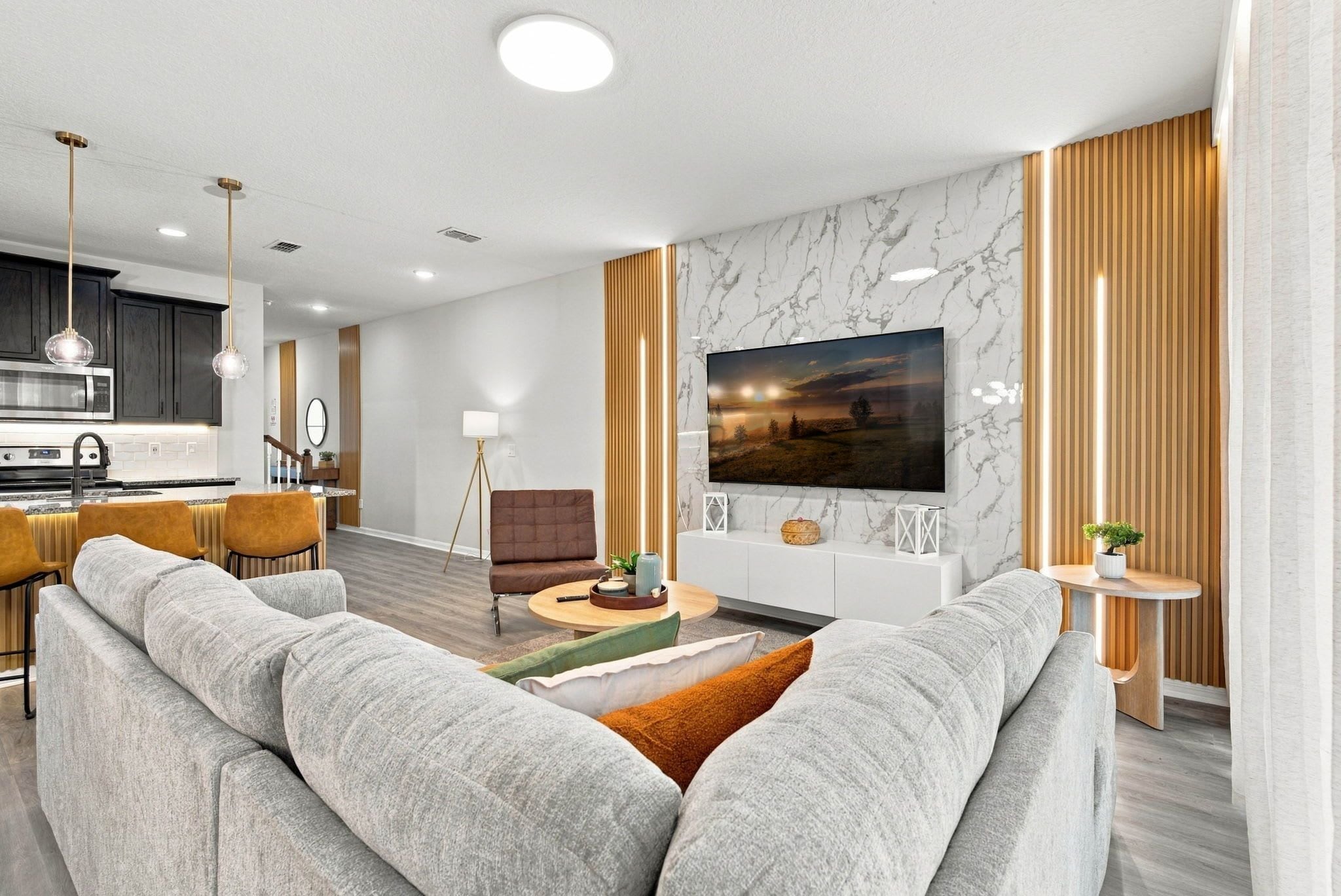 A modern living room with a large sofa, a flat-screen TV mounted on a marble wall, and a kitchen in the background.