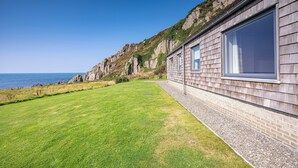 Un bâtiment situé sur la côte, doté de grandes fenêtres, d’une pelouse verdoyante et d’une falaise rocheuse.
