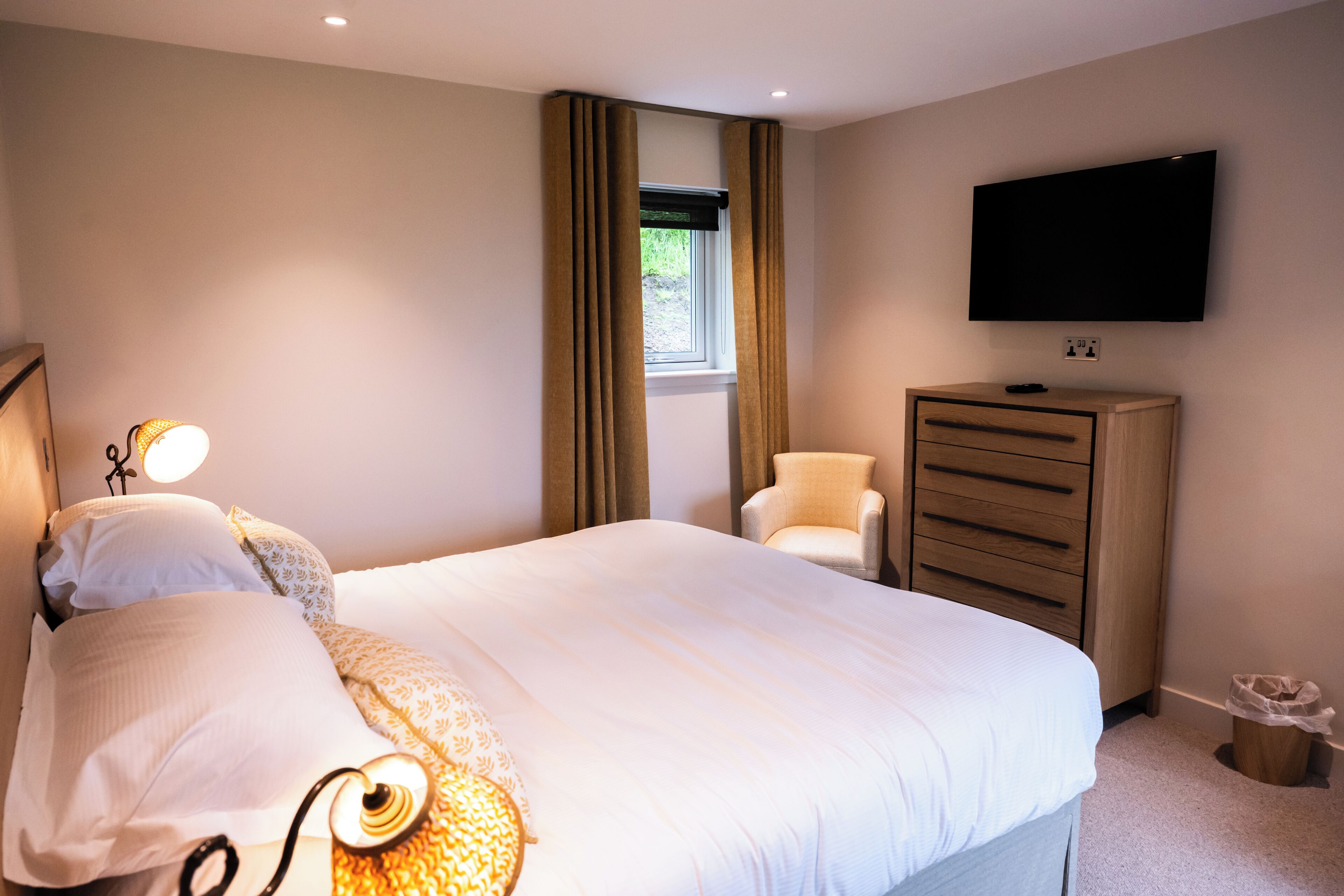 Une chambre d’hôtel avec un lit, un téléviseur fixé au mur, une commode, une chaise et une fenêtre avec des rideaux.