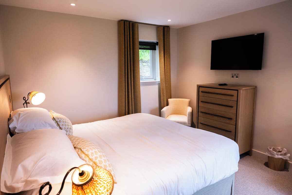 Une chambre d’hôtel avec un lit, un téléviseur fixé au mur, une commode, une chaise et une fenêtre avec des rideaux.