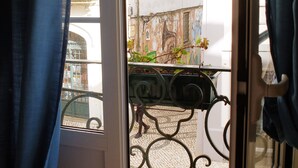 Vista a través de una puerta con reja metálica, un patio con baldosas y un edificio de fachada blanca y ventana.