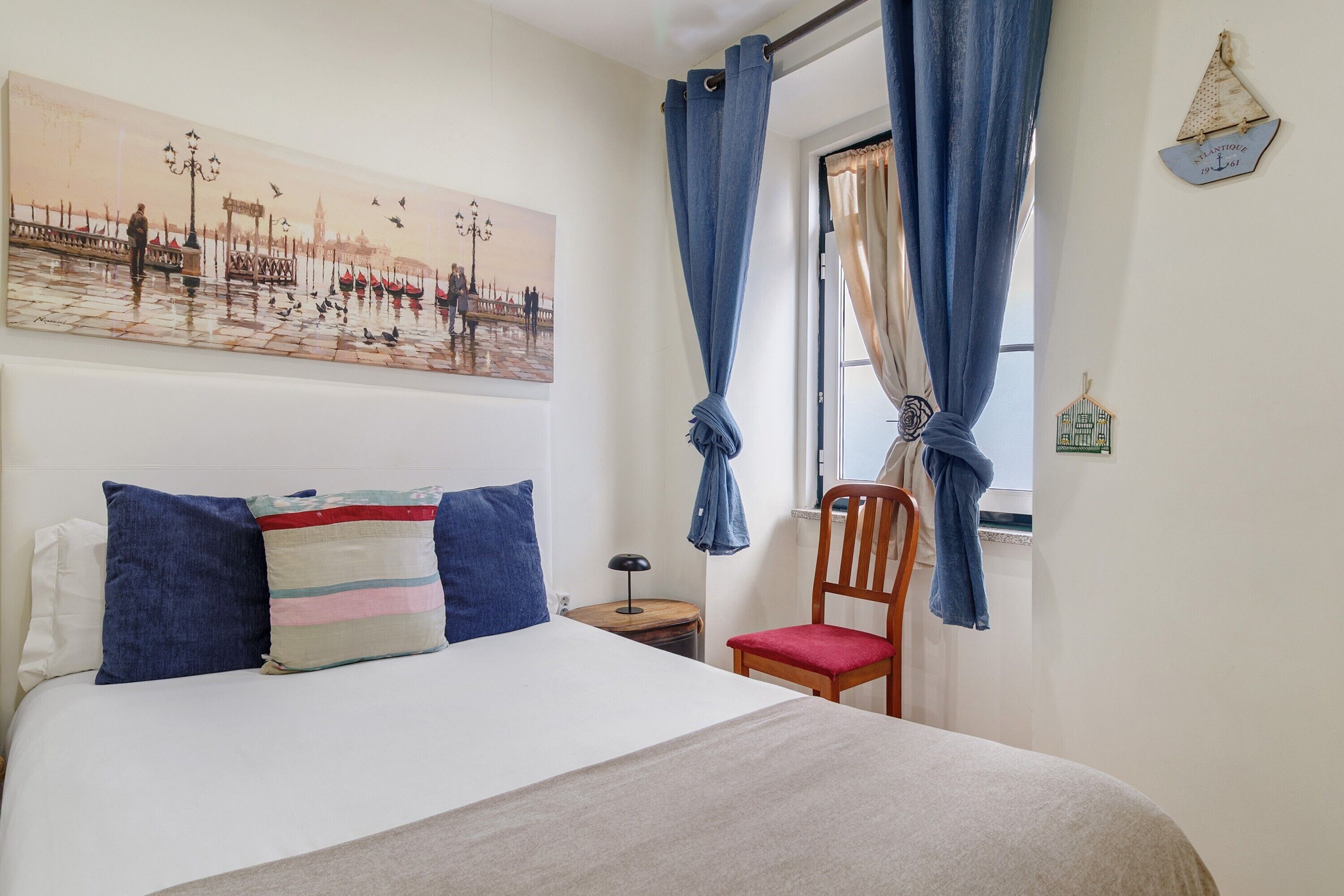A bedroom with a bed, a chair, a nightstand, a painting, and a window with curtains.