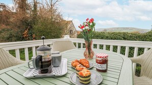 Une table dressée avec une cafetière, des tasses, des pâtisseries, un vase rempli de fleurs et un pot de confiture, le tout placé sur une terrasse avec des chaises.