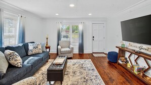 Una sala de estar con un sofá, un reposapiés y una mesa de centro. Se observa una silla, un mueble para televisión y una estantería.