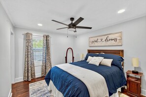 Ein Schlafzimmer mit Bett, Deckenventilator, Fenster mit gemusterten Vorhängen, Nachttisch mit Lampe und einer Wandtafel.