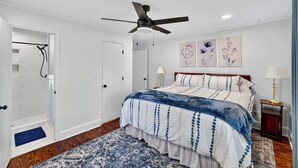 Un dormitorio con cama, ventilador de techo, acceso a una ducha y una obra de arte en la pared.