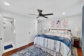 A bedroom with a bed, a ceiling fan, a door to a shower, and artwork on the wall.