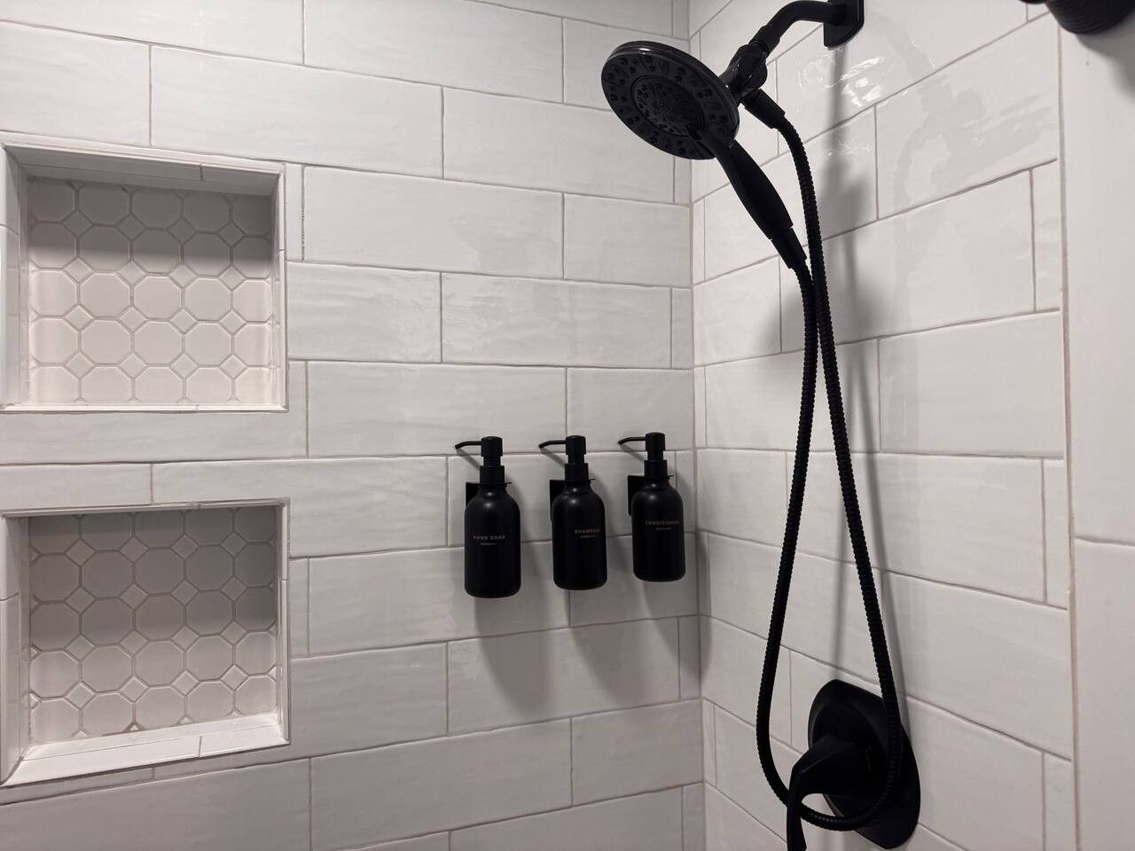 A shower area with a handheld showerhead, a wall-mounted showerhead, and three bottles of shampoo on the wall.
