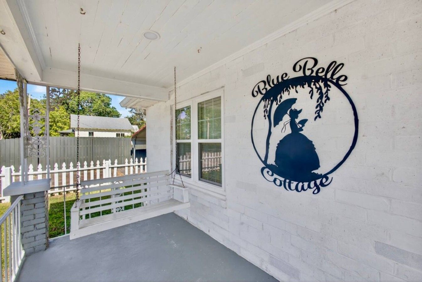 Eine Veranda mit einer Schaukel, einem weißen Zaun und einer Wand mit dem Logo von 'Blue Belle Cottage'.