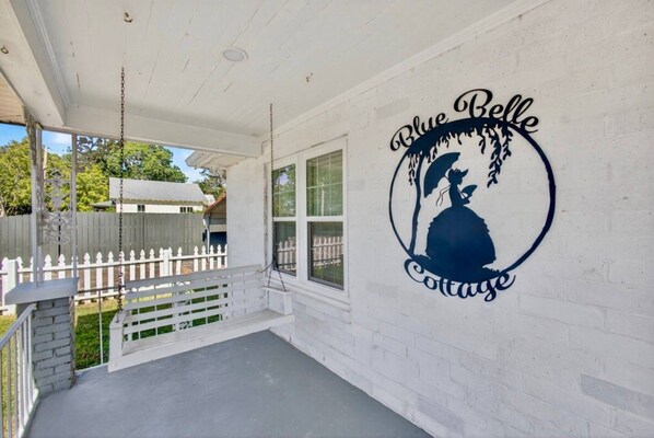 Un porxo amb un balancí, una tanca blanca i una paret amb el logotip de 'Blue Belle Cottage'.