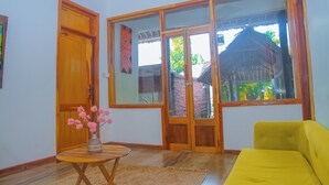 A room with a wooden door, a large window with a view of a traditional house, a yellow sofa, and a small table with a vase of flowers. - APAO VILLAGE & RESORT LODGE (Dar es Salaam)