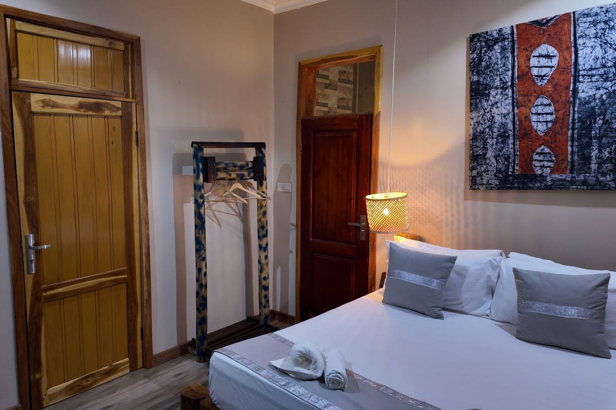 Ett hotellrum med en dörr av trä, en klädställning, en säng med kuddar, ett sängbord med en flaska och glas, och en väggmonterad lampa.
