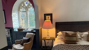 Une chambre avec un grand lit, une chaise, une table de chevet avec une lampe, une fenêtre avec des vitraux et un tableau encadré au mur.