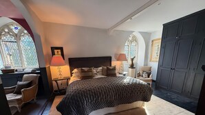Une chambre à coucher avec un grand lit, une chaise, une table de chevet, une lampe, une commode et une fenêtre ornée de vitraux.