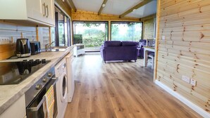 Une cuisine compacte équipée d’une cuisinière, d’un four et d’un lave-linge. Un canapé violet et une table à manger se trouvent dans l’espace de vie, et de grandes fenêtres offrent une vue sur la verdure environnante.