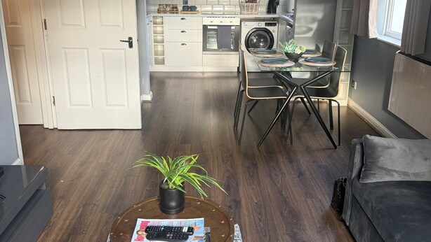 Ein modernes Wohnzimmer mit einer Couch, einem Couchtisch mit Zeitschriften, einem Essbereich mit einem Glastisch und einer Küche inklusive Waschmaschine.
