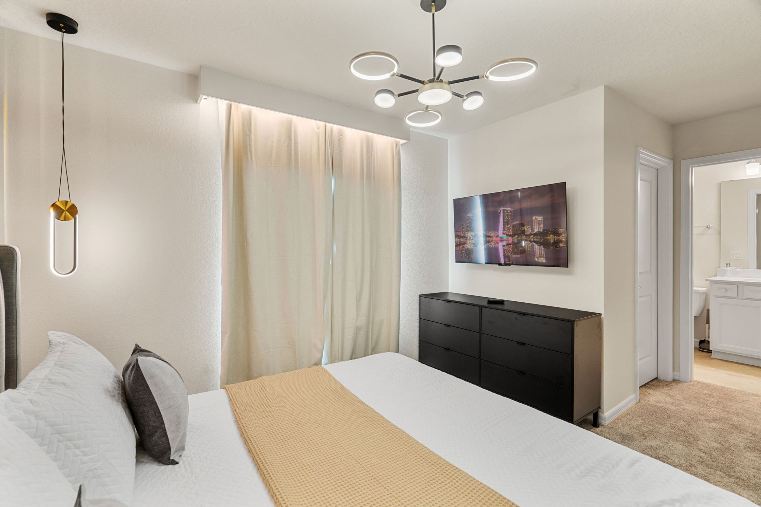 A modern bedroom with a large bed, a black dresser, a cityscape painting, and a bathroom visible through an open door.