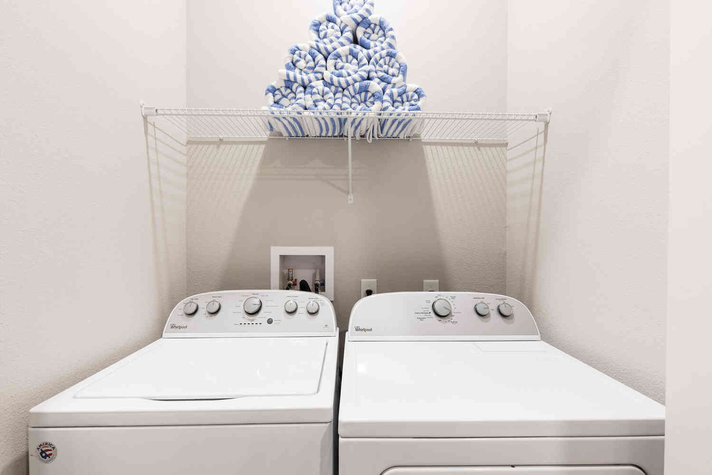 To hvide Whirlpool vaskemaskiner i et vaskerum med et metalstativ over dem.