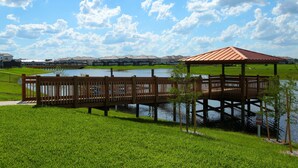 A wooden gazebo over a lake, surrounded by green grass and a row of houses in the background. - 4Bed Home in Storey Lake with Private Pool (Kissimmee)