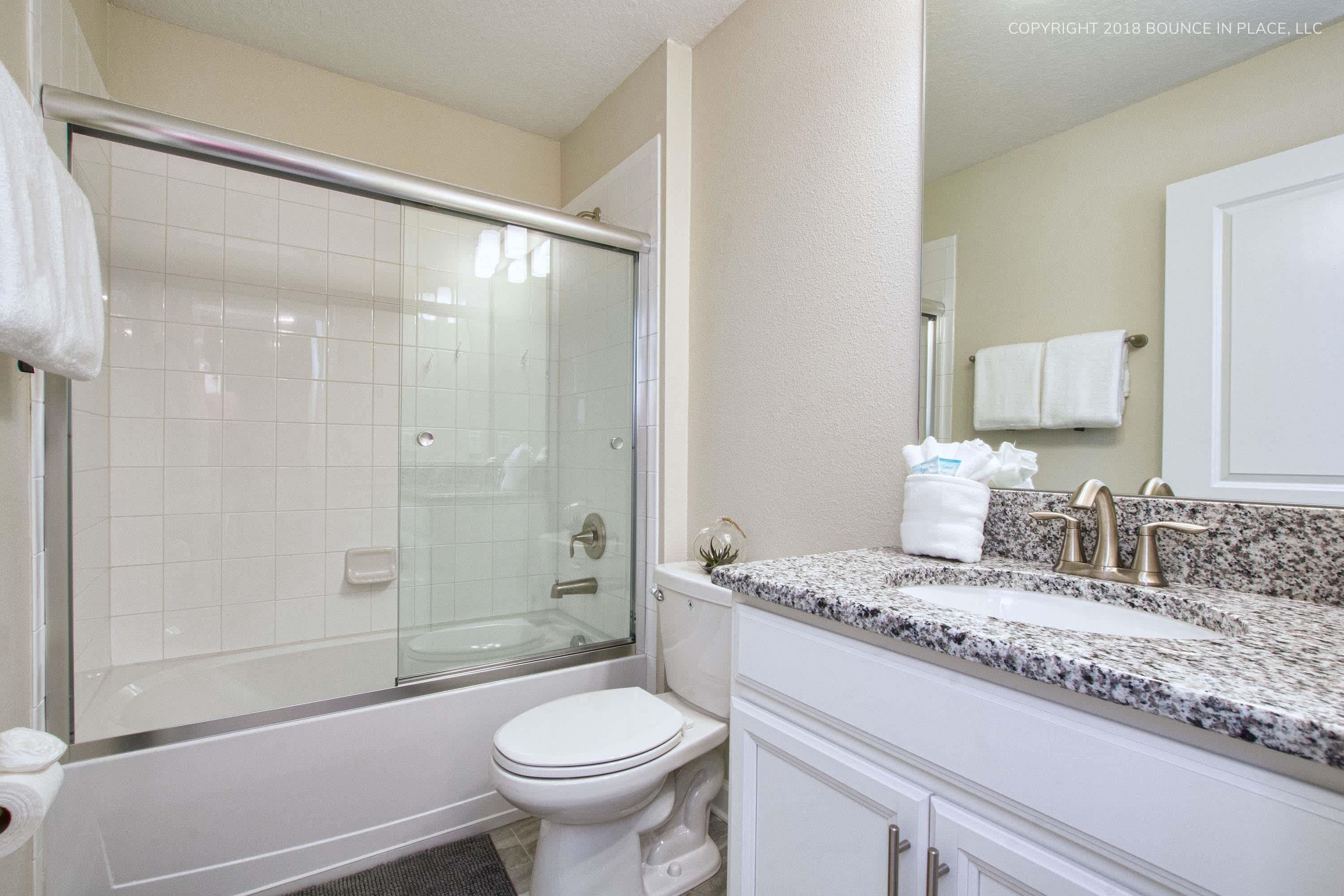 A bathroom with a glass-enclosed shower, a white toilet, and a granite countertop sink with a mirror above it.