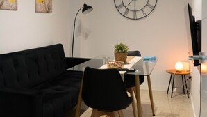 Ett modernt vardagsrum med en svart soffa, ett matbord med stolar, en väggklocka och en platt-TV.