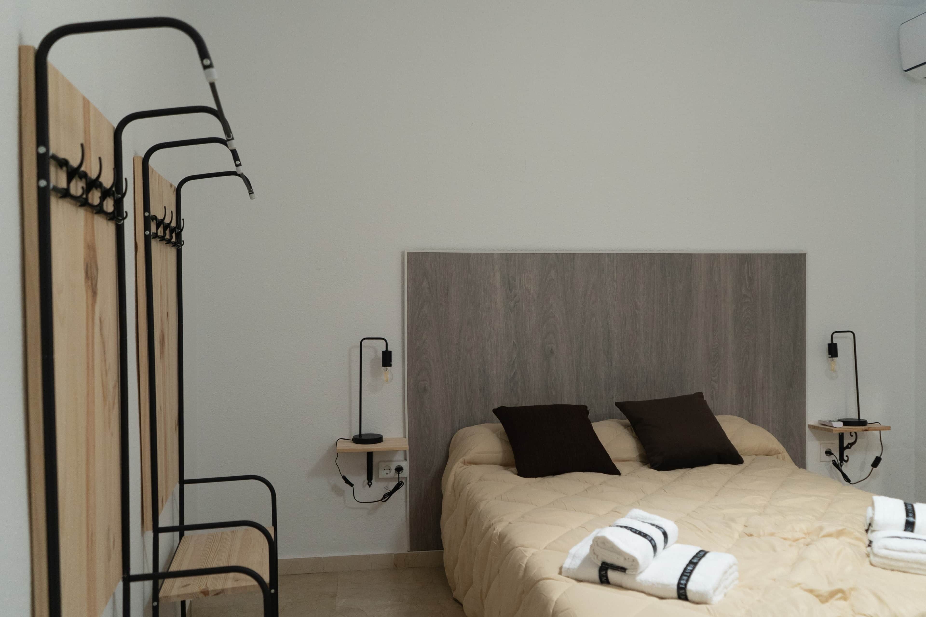 Ein Schlafzimmer mit einem Bett, zwei Kissen, einem Holzkopfstück, einem Nachttisch, einer Lampe und einem an der Wand befestigten Handtuchhalter.