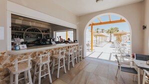 A bar area with stools, a counter with bottles, and a view of outdoor seating and palm trees. - Naxos Grand Hotel (Stelida)