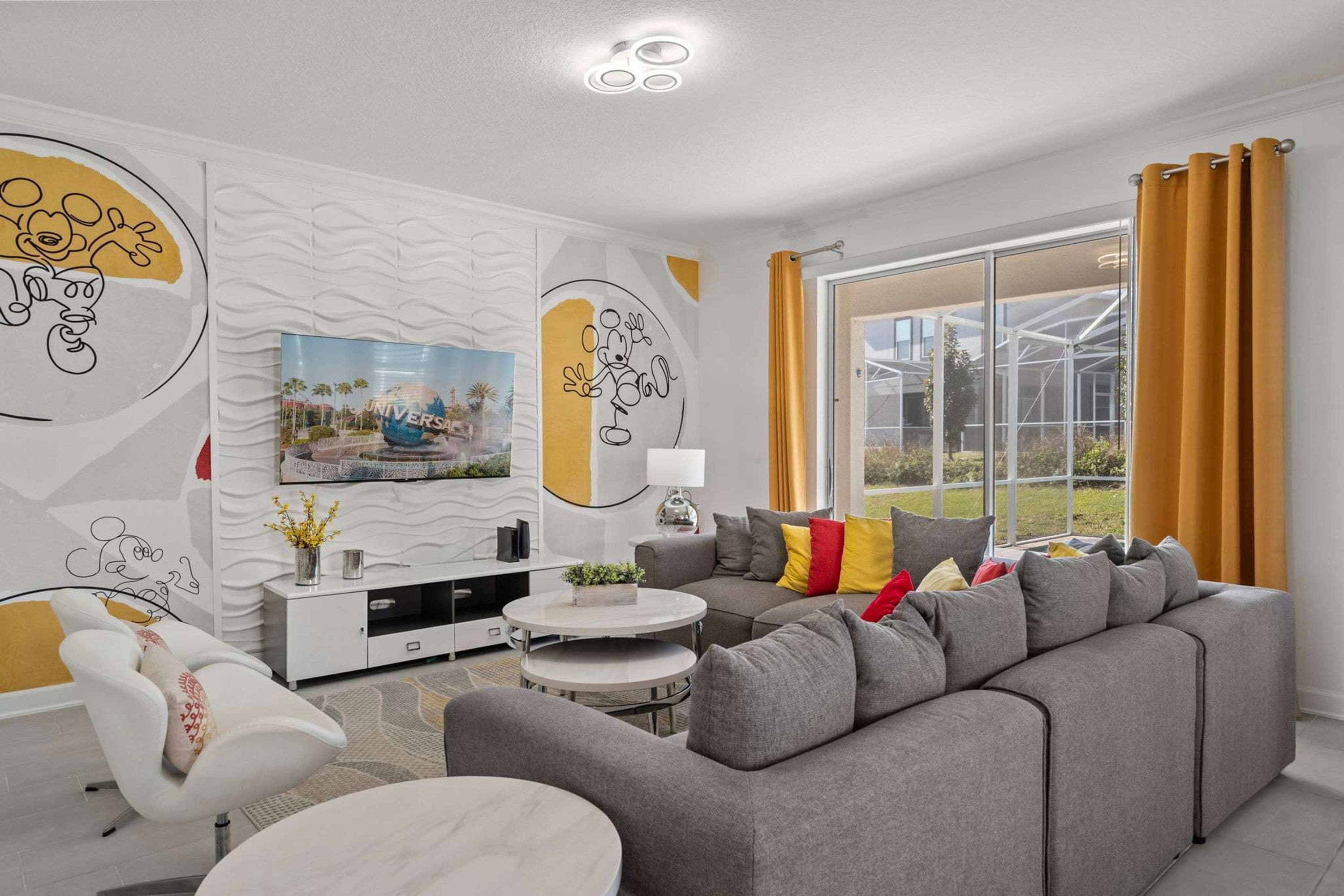 A modern living room with a grey sofa, a white armchair, and a round coffee table. There is a TV on a white cabinet, and a large window with curtains.