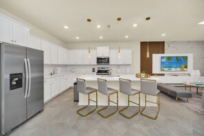 Una cocina moderna con gabinetes blancos, un refrigerador de acero inoxidable, una isla central con taburetes de bar y un televisor de pantalla plana montado en la pared.